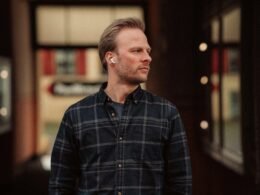 Ein Mann mit Ohrhörern steht vor einem modernen Gebäude und blickt zur Seite, urbanes Umfeld.