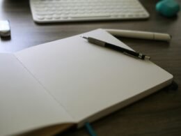 Ein geöffnetes Notizbuch mit einem Stift darauf liegt neben einer weißen Computertastatur auf einem Schreibtisch.
