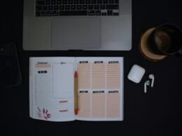 Ein offenes Notizbuch und eine Tasse Kaffee stehen auf einem ordentlich eingerichteten Schreibtisch in moderner Umgebung.