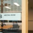Ein transparentes Schild mit der Aufschrift Meeting Room hängt an einer Glastür und kennzeichnet einen Besprechungsraum.