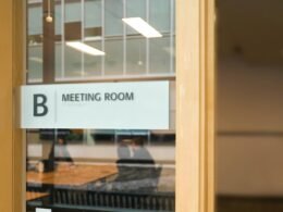 Ein transparentes Schild mit der Aufschrift Meeting Room hängt an einer Glastür und kennzeichnet einen Besprechungsraum.