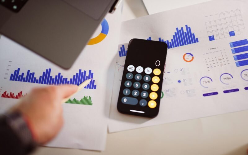 Taschenrechner auf einem Holztisch neben einem aufgeklappten Laptop, Fokus auf Arbeitsorganisation