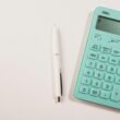 Ein türkisfarbener Taschenrechner neben einem weißen Stift auf weißem Hintergrund, geeignet für Themen wie Finanzen oder Online-Tools.