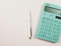 Ein türkisfarbener Taschenrechner neben einem weißen Stift auf weißem Hintergrund, geeignet für Themen wie Finanzen oder Online-Tools.