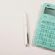Ein türkisfarbener Taschenrechner neben einem weißen Stift auf weißem Hintergrund, geeignet für Themen wie Finanzen oder Online-Tools.
