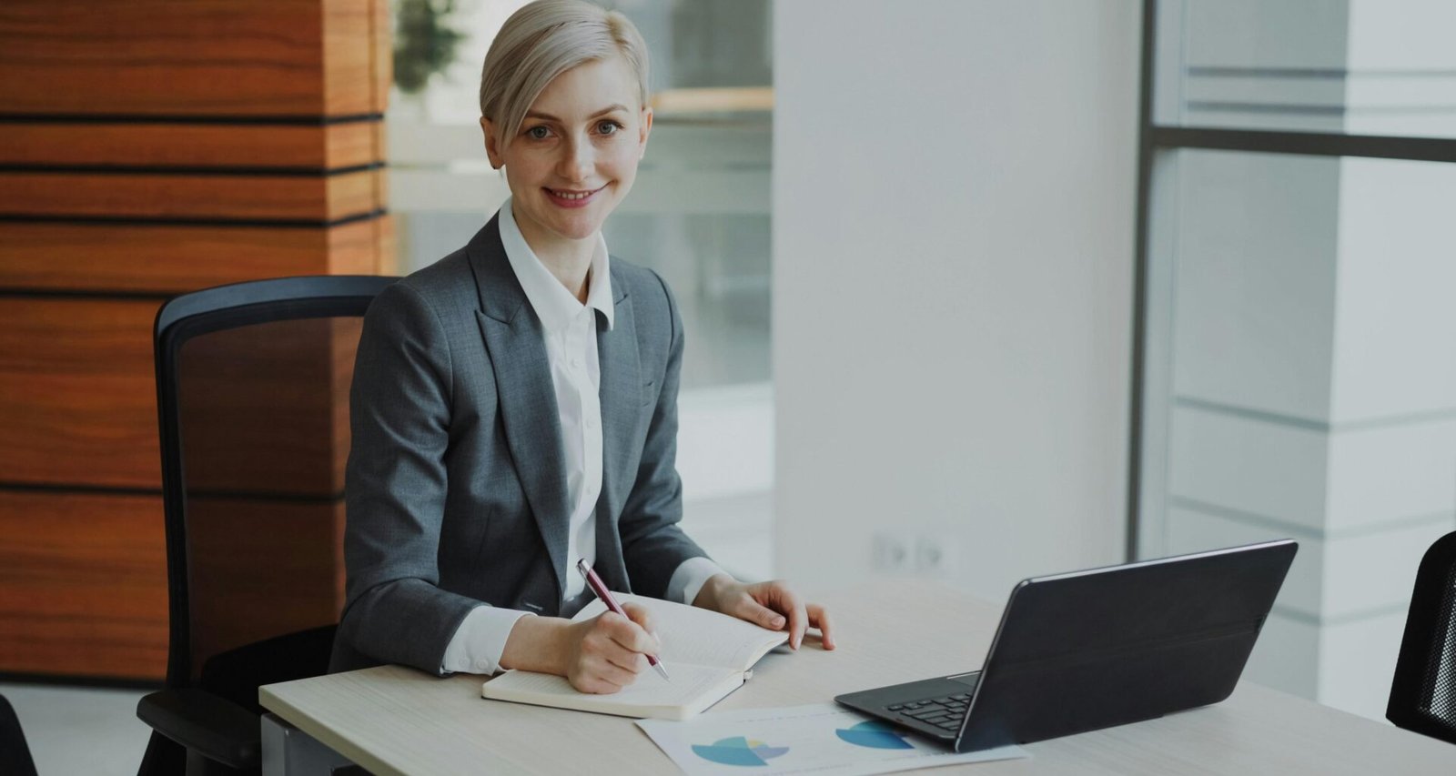 Eine Frau im Business-Anzug sitzt an einem Schreibtisch und arbeitet konzentriert an ihren Unterlagen im Büro.