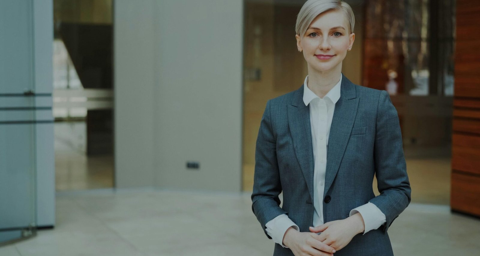 Eine berufstätige Frau im Anzug steht lächelnd und selbstsicher vor neutralem Hintergrund