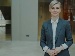 Eine berufstätige Frau im Anzug steht lächelnd und selbstsicher vor neutralem Hintergrund