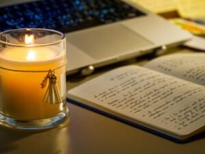 Beleuchtete Kerze steht neben einem offenen Notizbuch und einem aufgeklappten Laptop auf einem Arbeitstisch.