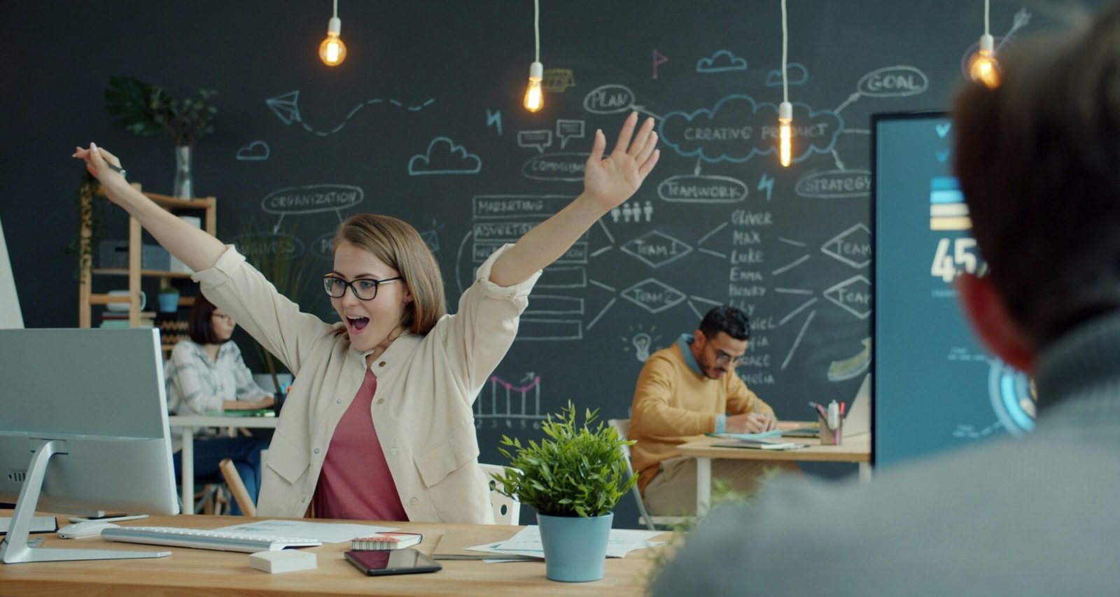 Frau sitzt an ihrem Büroschreibtisch und hebt jubelnd die Arme für ein erfolgreiches Arbeitsergebnis