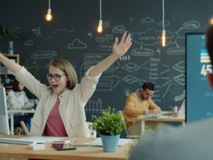 Frau sitzt an ihrem Büroschreibtisch und hebt jubelnd die Arme für ein erfolgreiches Arbeitsergebnis