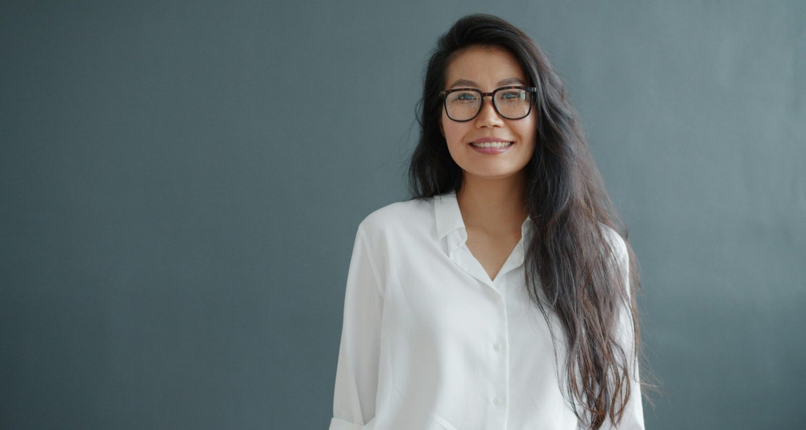 Eine junge Frau mit Brille und langen Haaren lächelt freundlich in die Kamera, Hintergrund ist einfarbig grau