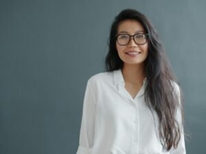 Eine junge Frau mit Brille und langen Haaren lächelt freundlich in die Kamera, Hintergrund ist einfarbig grau