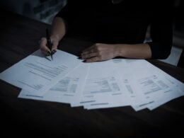 Frau sitzt an einem Tisch mit zahlreichen Papieren und arbeitet an Dokumenten, Schreibtisch voll Unterlagen