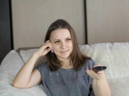 Frau sitzt auf einem Sofa und hält eine Fernbedienung, gemütliche Alltagsatmosphäre im Wohnzimmer