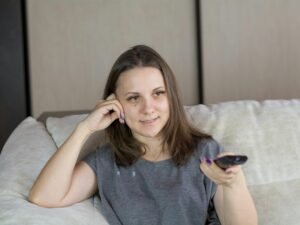 Frau sitzt auf einem Sofa und hält eine Fernbedienung, gemütliche Alltagsatmosphäre im Wohnzimmer