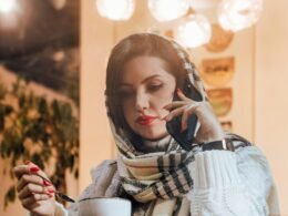 Frau hält eine Tasse Kaffee und spricht dabei unterwegs in ein Mobiltelefon, dargestellt in Alltagsszenerie