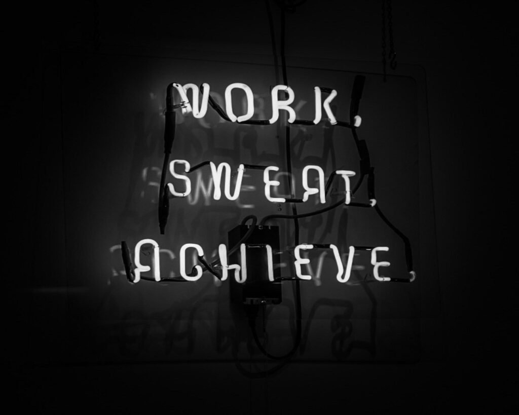 Leuchtendes Neon-Schild mit der Aufschrift Work, Sweat, Achieve vor dunklem Hintergrund