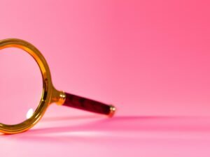 Eine einzelne Lupe liegt zentral auf einem einfarbig rosa Hintergrund und symbolisiert präzise Analyse.