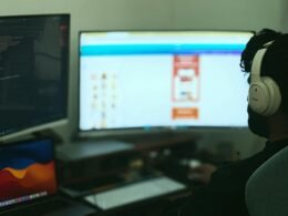Person arbeitet an einem Schreibtisch mit mehreren Monitoren und Computer, umgeben von Technik im modernen Büro.