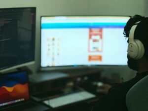 Person arbeitet an einem Schreibtisch mit mehreren Monitoren und Computer, umgeben von Technik im modernen Büro.