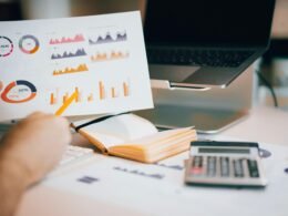 Person hält ein Blatt Papier über ein aufgeklapptes Laptop auf einem Schreibtisch im Büro