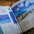 Eine Person hält ein geöffnetes Buch, auf dessen Seite ein Foto eines Flugzeugs zu sehen ist, Fokus auf Reisen und Technik