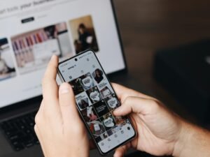 Eine Person hält ein Smartphone in der Hand und sitzt vor einem aufgeklappten Laptop auf einem Schreibtisch.