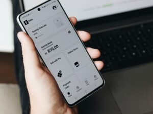 Eine Person hält ein Smartphone in der Hand und sitzt vor einem aufgeklappten Laptop, beide Geräte werden gleichzeitig verwendet.