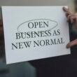Eine Person hält ein Schild mit der Aufschrift 'Open Business as New Normal' in den Händen vor sich in einem urbanen Umfeld