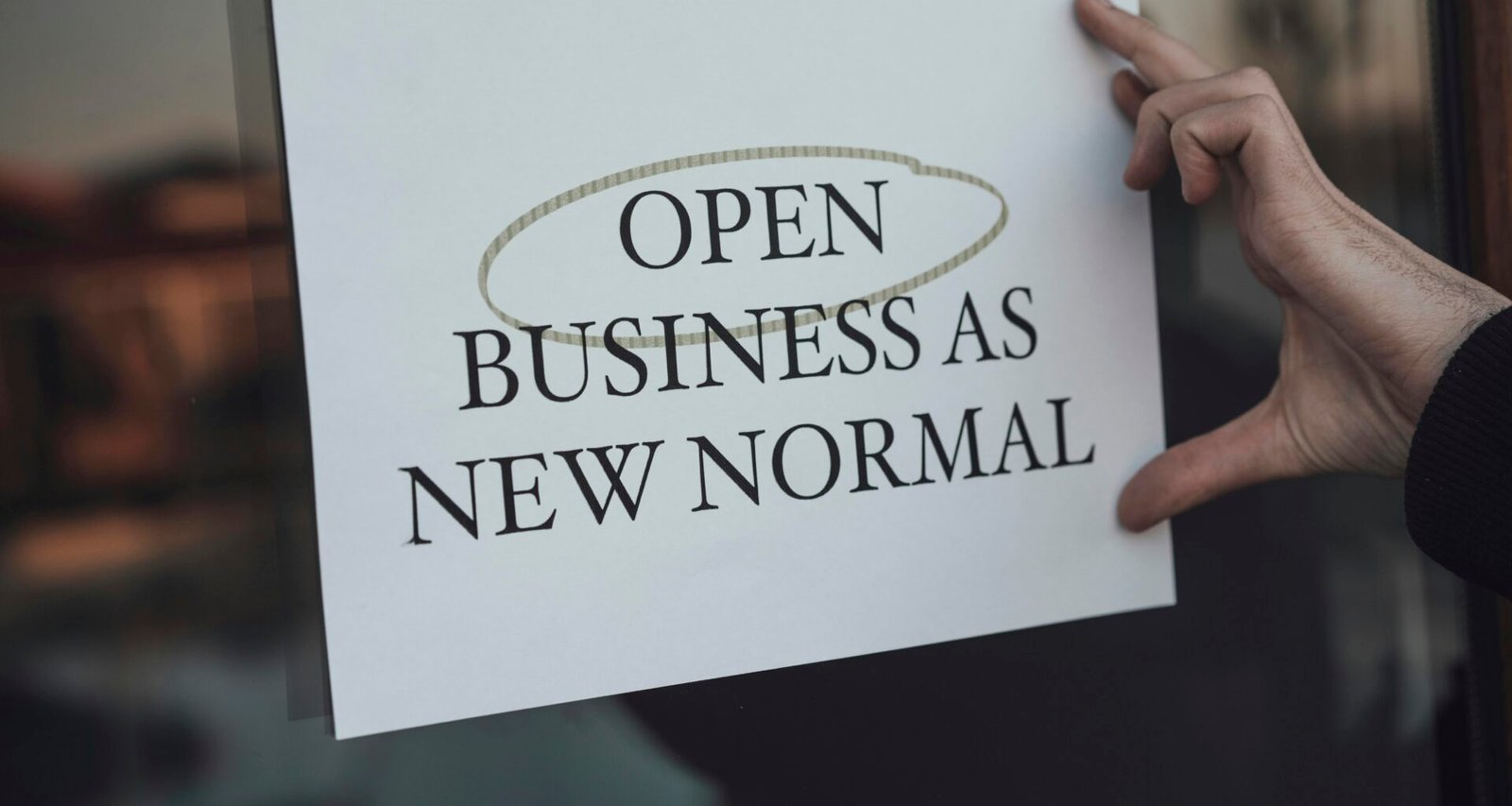 Eine Person hält ein Schild mit der Aufschrift 'Open Business as New Normal' in den Händen vor sich in einem urbanen Umfeld