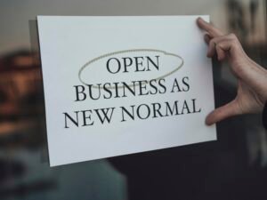 Eine Person hält ein Schild mit der Aufschrift 'Open Business as New Normal' in den Händen vor sich in einem urbanen Umfeld
