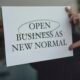Eine Person hält ein Schild mit der Aufschrift 'Open Business as New Normal' in den Händen vor sich in einem urbanen Umfeld