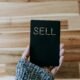 Person hält ein schwarzes Buch mit dem Wort 'Sell' nach oben – Symbolbild für Verkauf und Marketing