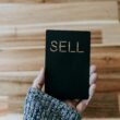 Person hält ein schwarzes Buch mit der Aufschrift 'Sell' in der Hand, geeignet für Artikel über Online-Vertrieb oder Verkaufsstrategien.