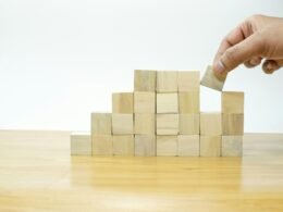 Person platziert einen Holzblock präzise auf einem Stapel, Symbol für Planung und Teambuilding im digitalen Arbeitsumfeld.
