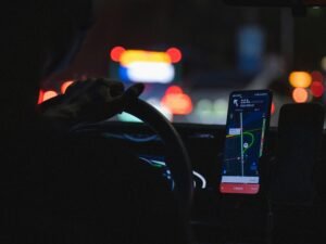 Eine Person fährt nachts ein Auto und hält dabei ein Smartphone in der Hand, Straße und Armaturenbrett sind beleuchtet.