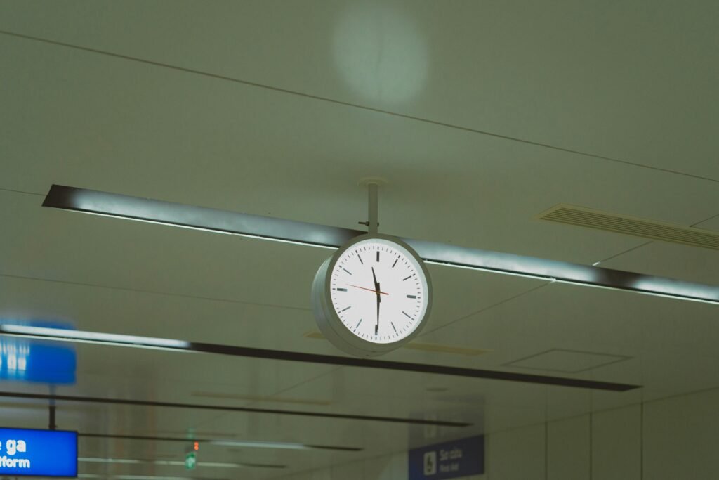 Große runde Uhr hängt von der Decke in einem modernen Flughafengebäude mit Glasdach und Menschen im Hintergrund.