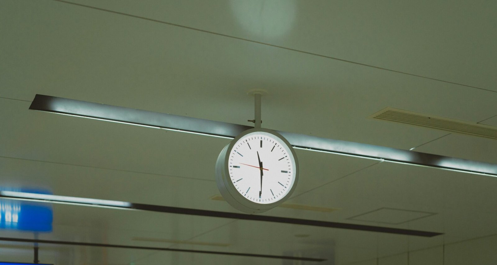 Große runde Uhr hängt von der Decke in einem modernen Flughafengebäude mit Glasdach und Menschen im Hintergrund.