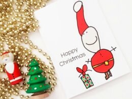 Eine Weihnachtskarte liegt auf einem Tisch neben einem geschmückten Weihnachtsbaum mit Lichtern und Kugeln.