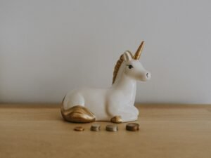 Weiß-goldene Keramik-Einhornfigur neben verstreuten Münzen, Aufsicht, passend zu Themen wie Online-Marketing und Finanzen.