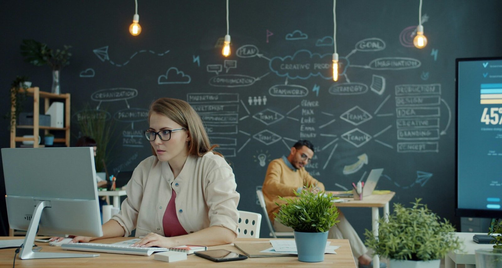 Eine Frau arbeitet an einem Computer in einem modernen Büro mit großer Tafel im Hintergrund.
