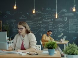 Eine Frau arbeitet an einem Computer in einem modernen Büro mit großer Tafel im Hintergrund.