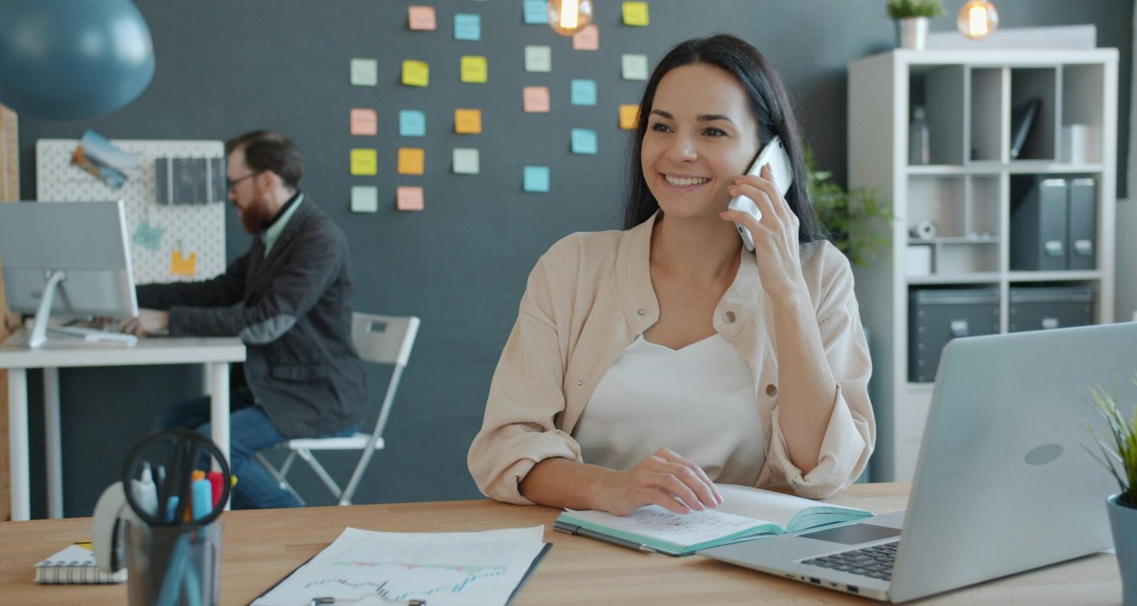 Frau sitzt am Schreibtisch, spricht am Telefon und nutzt einen Laptop, typische Arbeitsszene im modernen Büroalltag