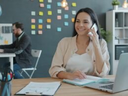 Frau sitzt am Schreibtisch, spricht am Telefon und nutzt einen Laptop, typische Arbeitsszene im modernen Büroalltag