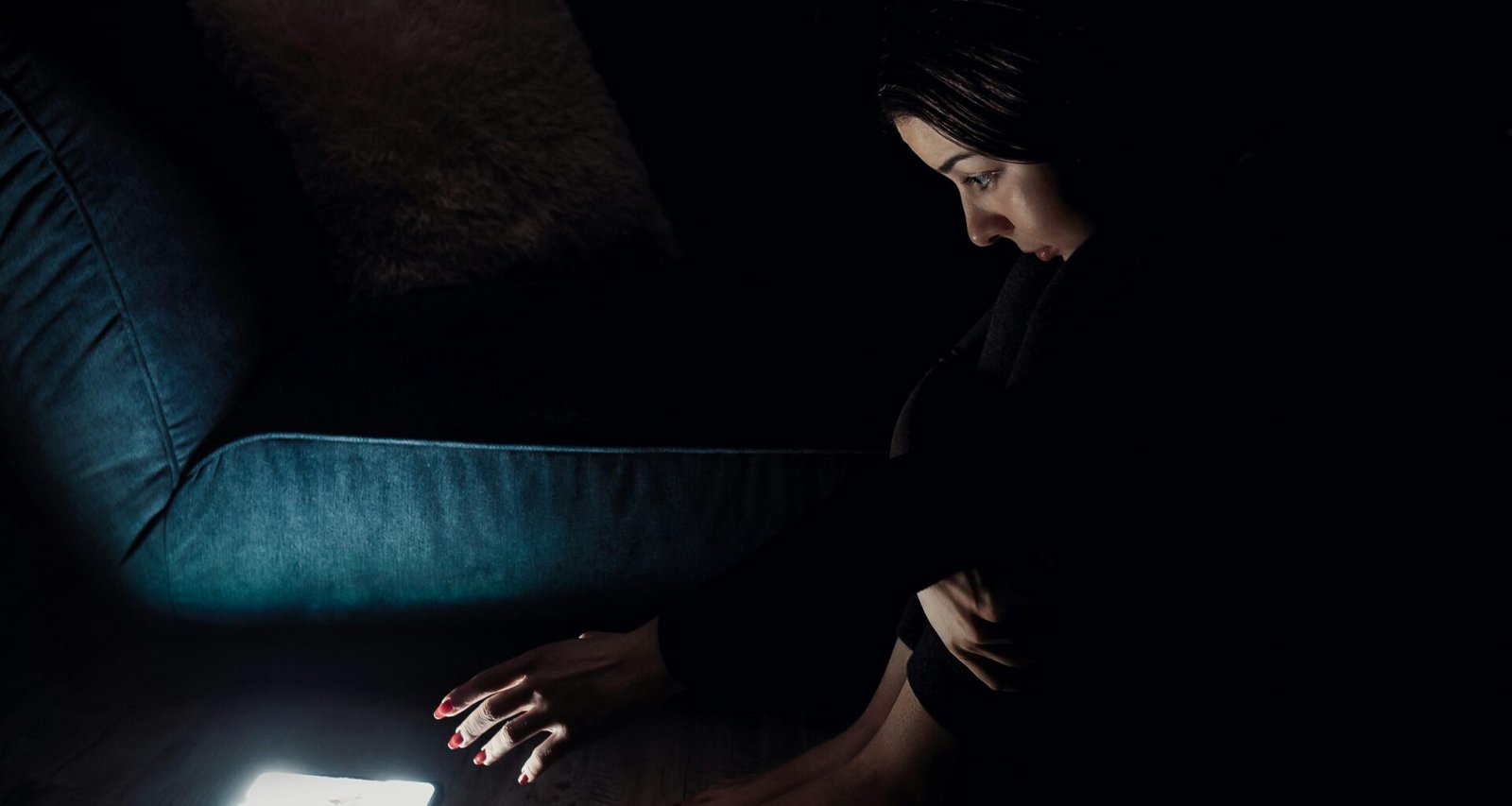 Frau mit schwarzem Langarmshirt sitzt auf blauem Sofa und nutzt moderne Technologien, geeignet für Artikel über Homeoffice oder Remote Work