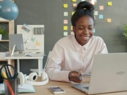 Frau arbeitet an einem Laptop am Schreibtisch, umgeben von modernen Arbeitsutensilien in einem hellen Büro.
