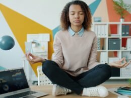 Frau meditiert entspannt am Schreibtisch vor einem Laptop in einer modernen Homeoffice-Umgebung
