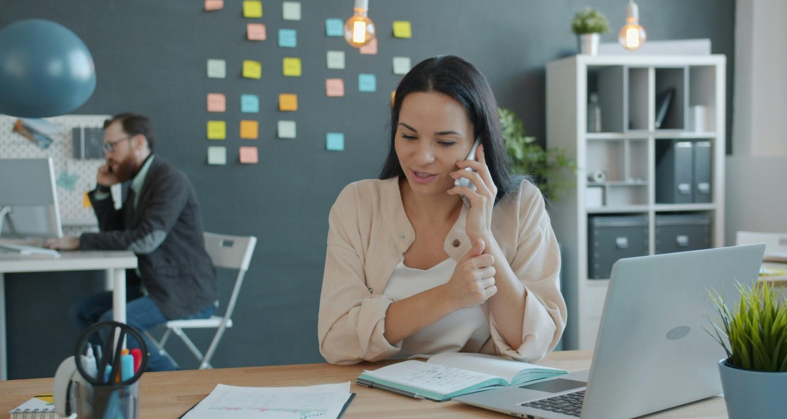 Frau sitzt am Schreibtisch, telefoniert und nutzt einen Laptop in einem modernen Arbeitsumfeld