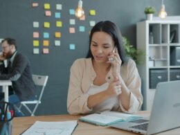 Frau sitzt am Schreibtisch, telefoniert und nutzt einen Laptop in einem modernen Arbeitsumfeld